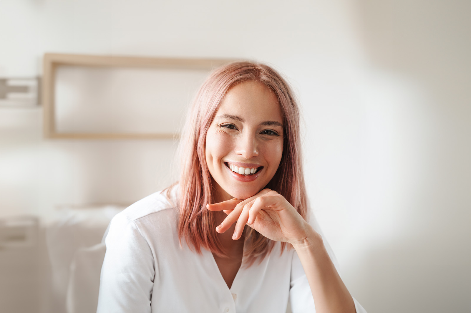 The image shows a smiling woman with pink hair, wearing a white top, seated with her chin resting on her hand, looking directly at the camera.