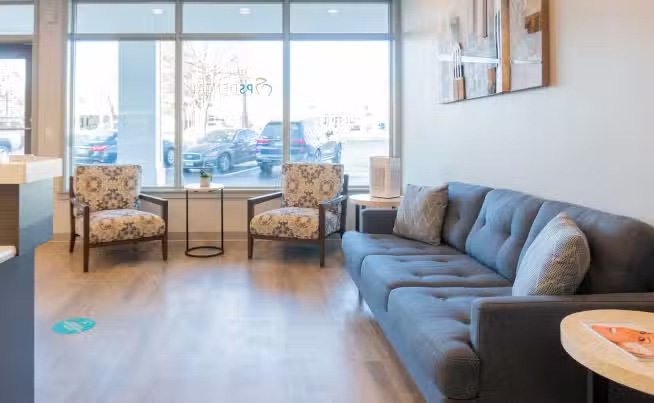 The image shows an interior view of a modern living room with a couch, chairs, a coffee table, and a large window allowing natural light into the space.