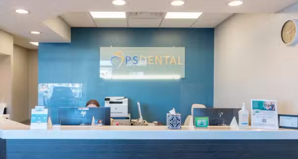 The image shows an interior view of a dental clinic reception area with a receptionist seated behind a counter, featuring a blue wall with a logo and text, a waiting area with chairs, and a professional atmosphere.