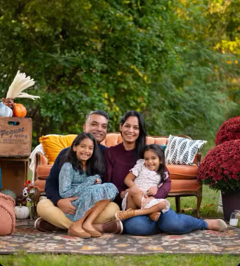 The image depicts a family of four sitting on an outdoor rug with a festive fall setting in the background, including autumn leaves and pumpkins, as well as a table with decorative items.