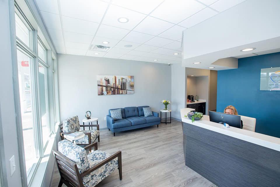 The image shows an interior space that appears to be a modern waiting room or reception area with a clean, bright design featuring light-colored walls, a large window allowing natural light, blue accent walls, white flooring, and contemporary furniture including sofas and chairs.