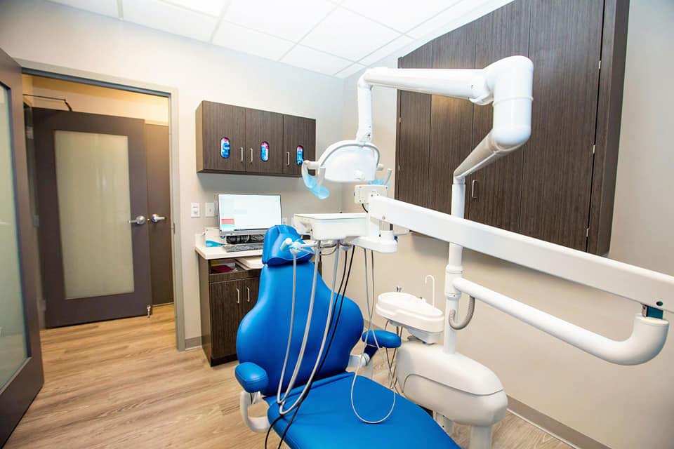 The image depicts a dental office interior with a blue dental chair, a white dental light, and various dental equipment visible behind a counter with a computer monitor on top.