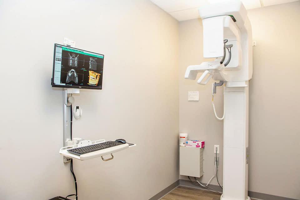 The image shows a medical setting with an X-ray machine and a monitor displaying an X-ray image, set up in a room with a computer desk and a chair.