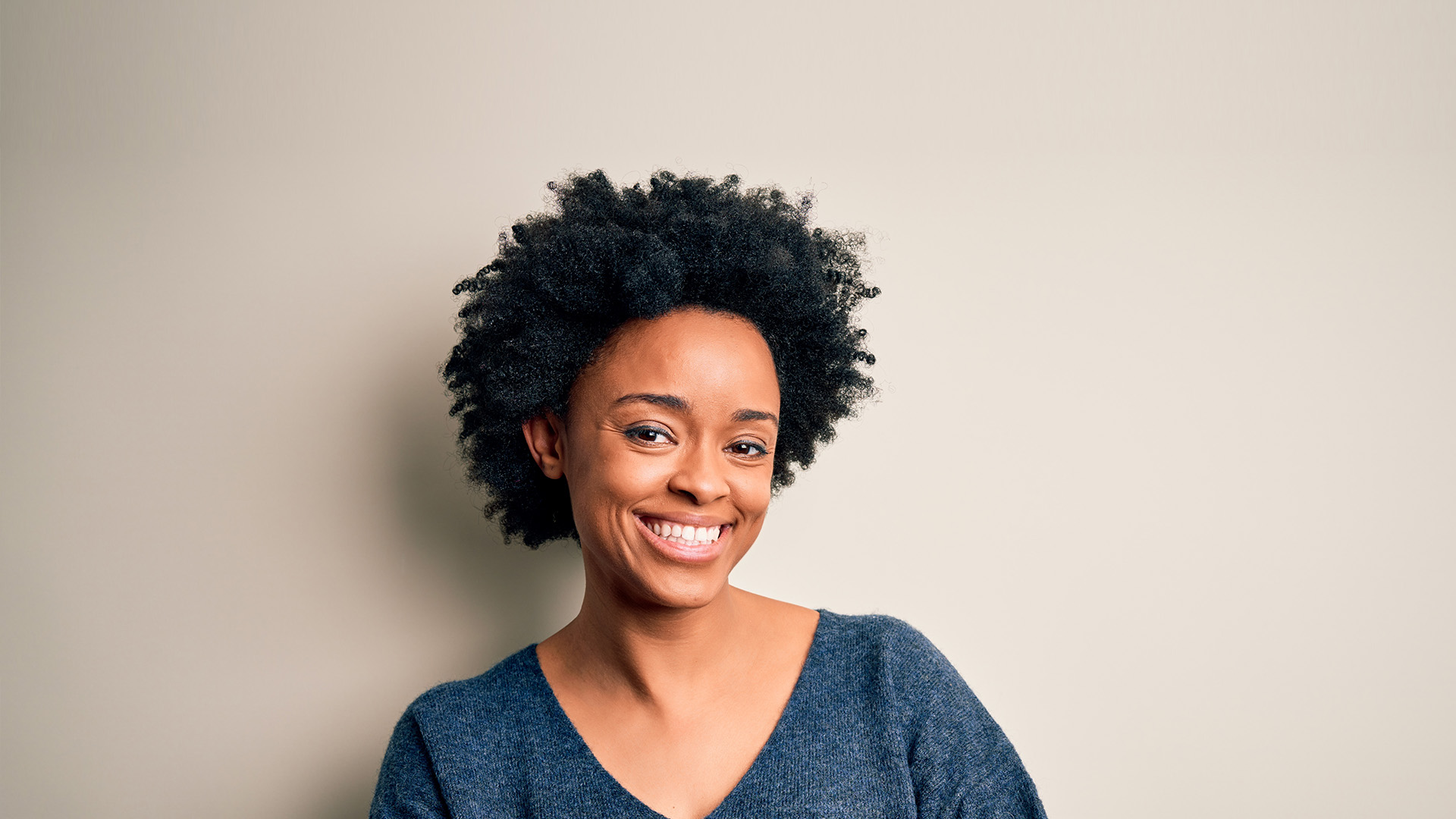 The image features a smiling woman with curly hair leaning against a light background, wearing a dark top.