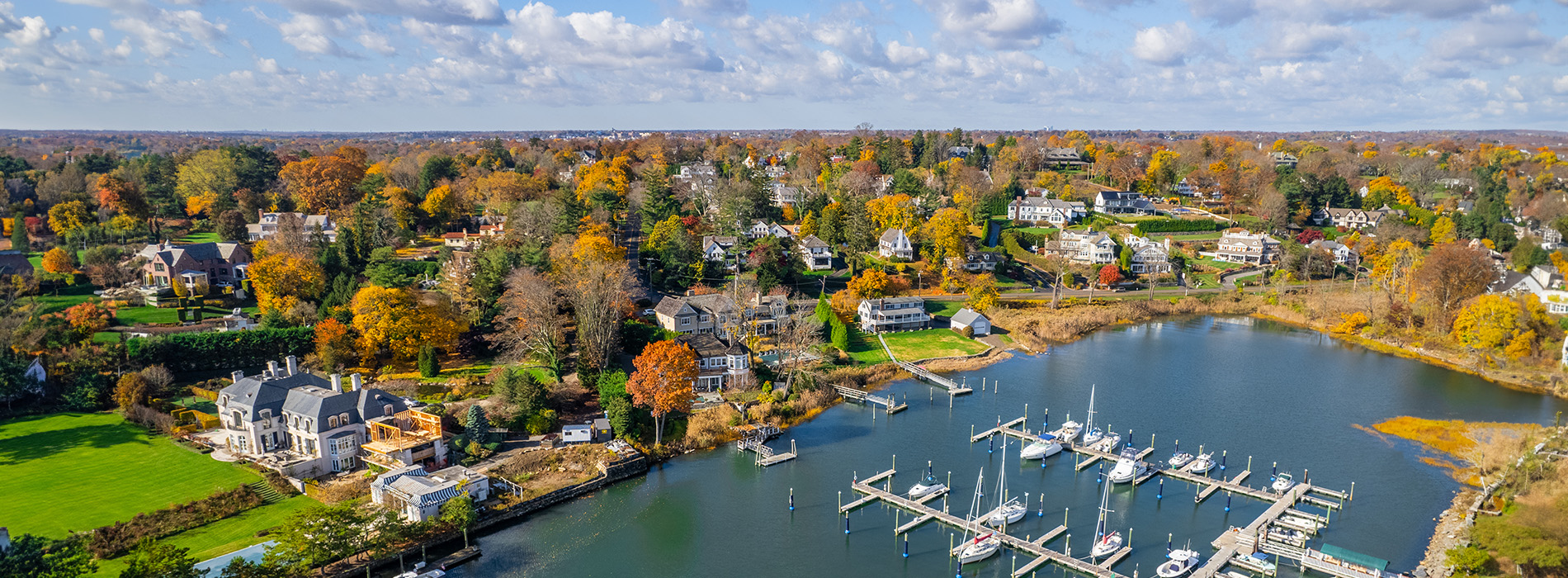 The image depicts a serene suburban landscape with a large body of water, docked boats, a marina, a residential area with houses, trees with autumn foliage, and clear skies above.