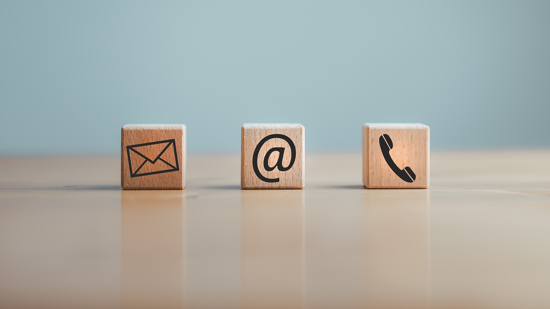 The image shows four wooden blocks with symbols for email, phone, and mailbox on them arranged in a line, with an enlarged photograph of an email icon superimposed at the end, all set against a blurred background that suggests a tabletop setting.