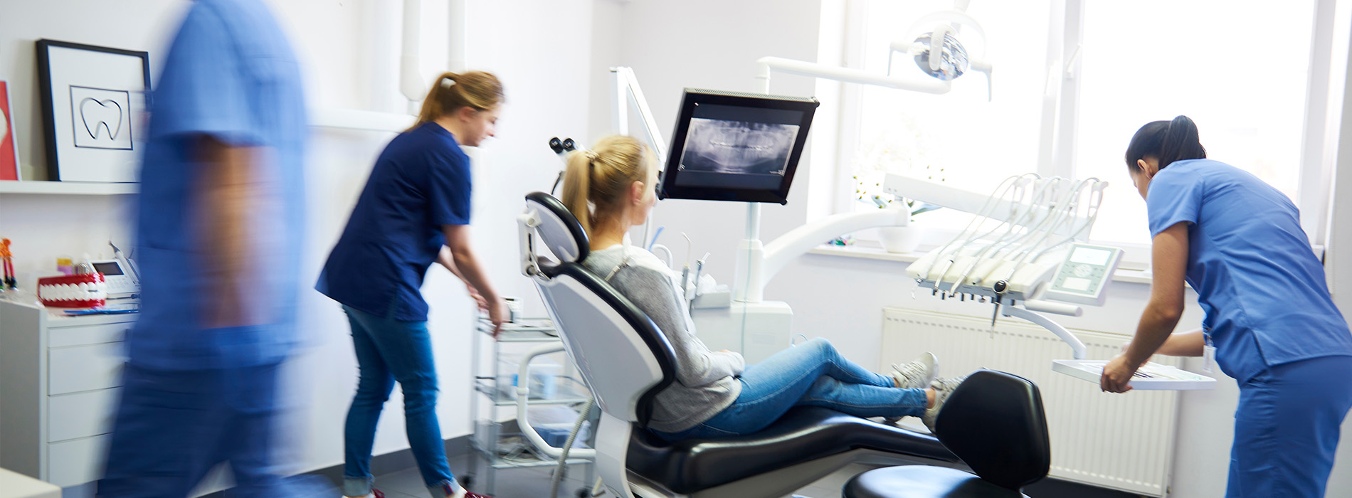 A photograph shows two women in scrubs attending to a patient s dental chair, with a blurred background depicting a medical office setting.