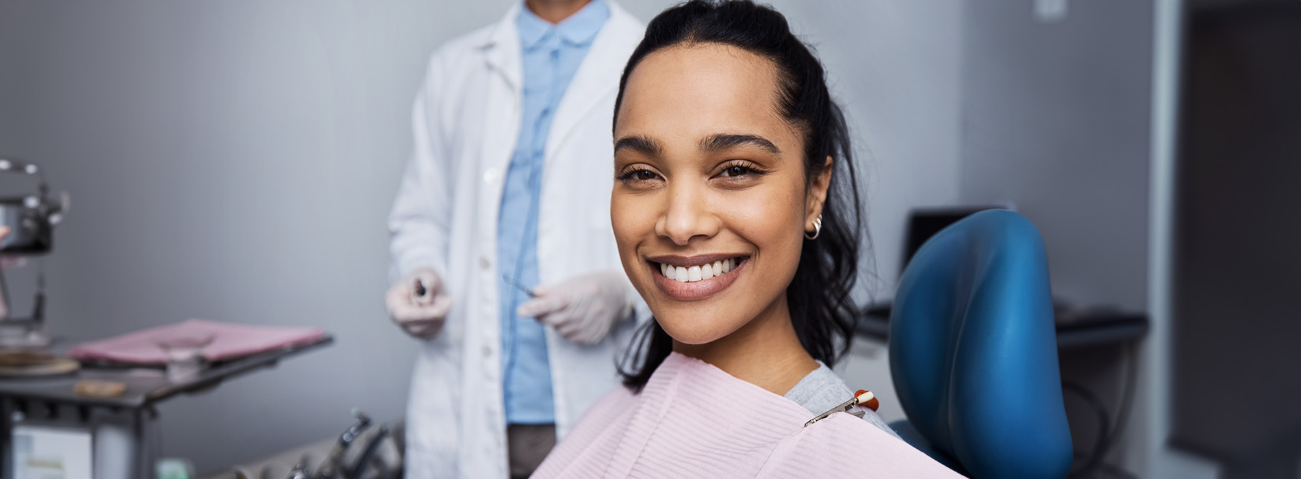 A smiling woman sitting in a dental chair with a dentist standing behind her, both wearing face masks.