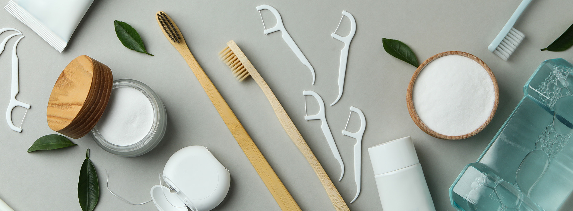 A collection of eco-friendly personal care products displayed on a gray surface with green leaves and wooden toothbrushes.