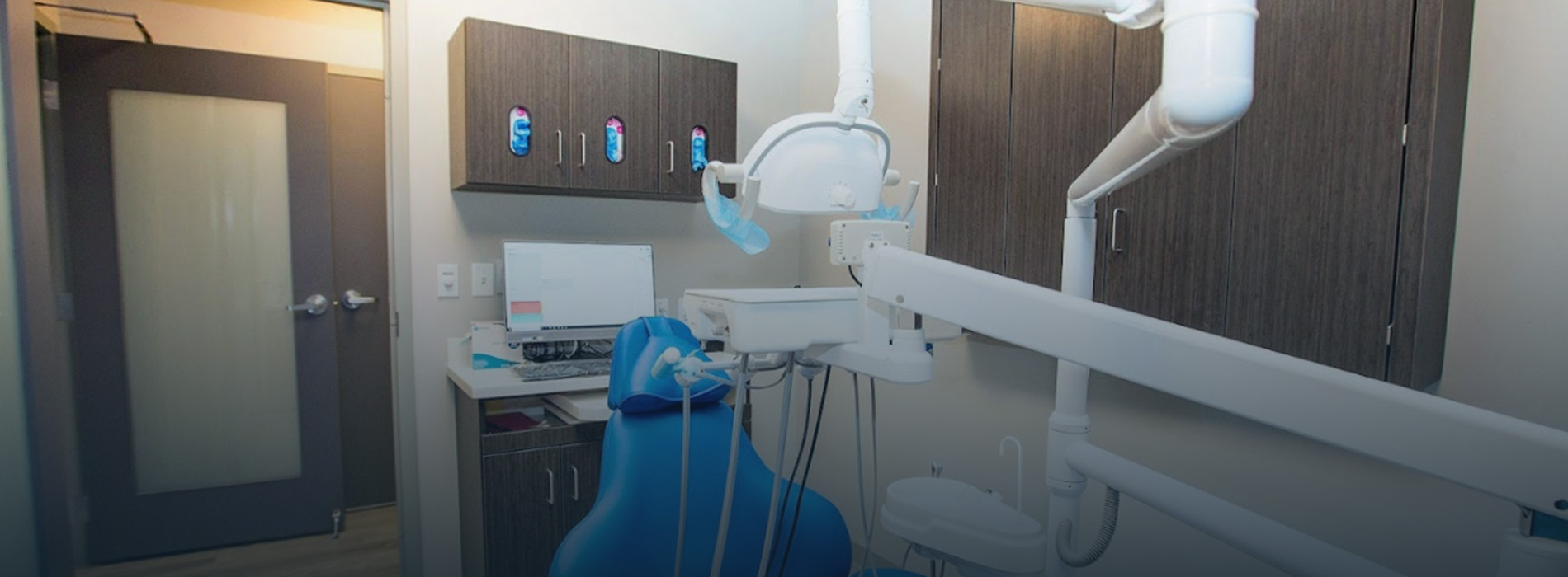 The image shows a modern dental office interior with various equipment and a blue chair, viewed from an angle that includes a large window allowing natural light into the room.