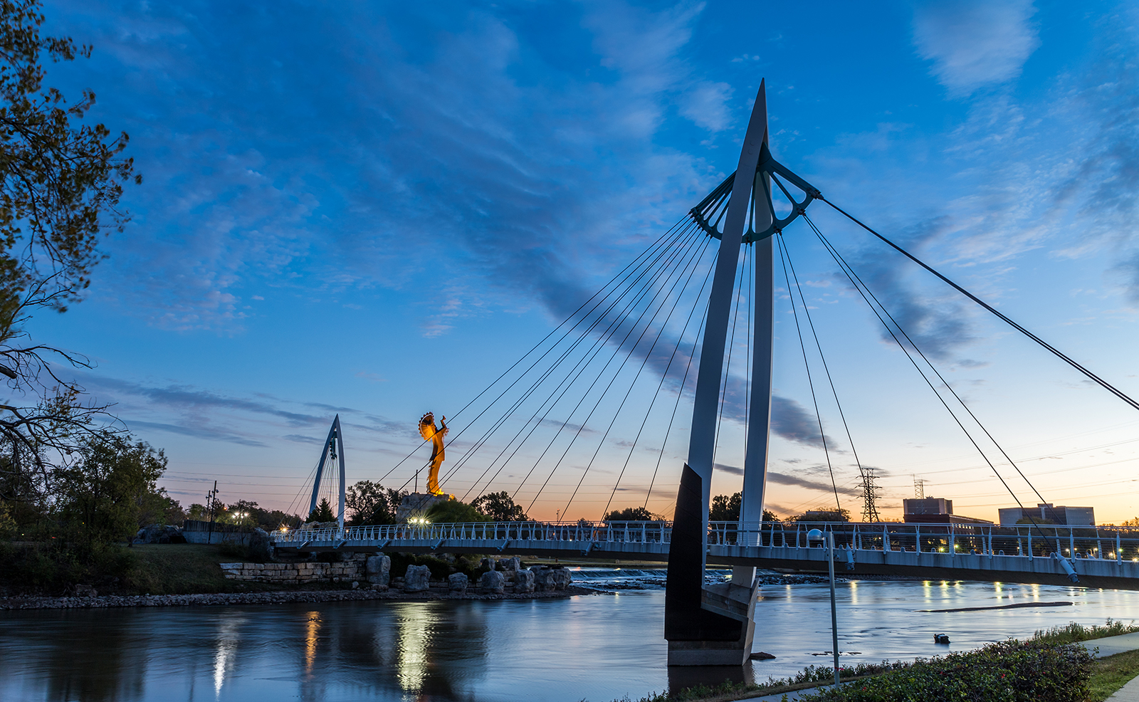 The image depicts a scenic view of a bridge at dusk with a city skyline in the background, featuring a suspension bridge with a distinctive design and a river running beneath it, illuminated by streetlights and the glow of the setting sun.
