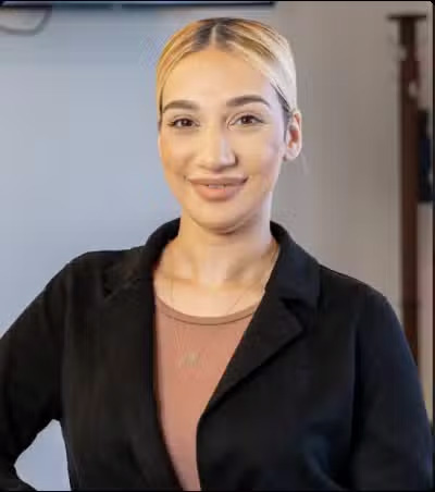 The image shows a person standing with a confident posture, smiling slightly, wearing a black blazer over a top with a V-neckline, and sporting short hair.