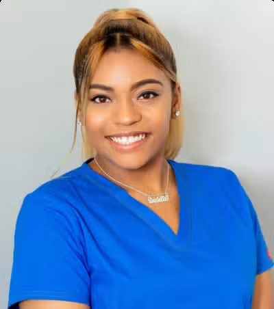 The image features a person smiling at the camera, wearing a blue shirt and a name tag with a necklace around their neck. They appear to be a healthcare professional, possibly a nurse or a doctor, given the attire and setting.