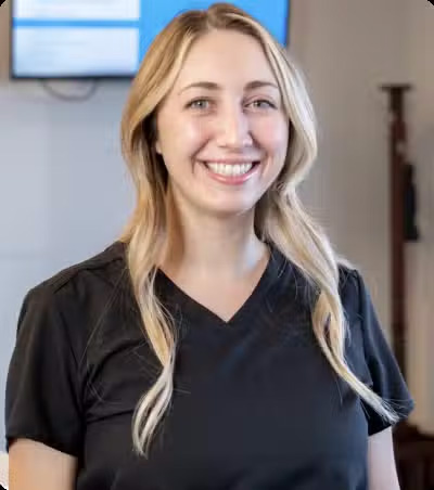 The image shows a woman with blonde hair wearing a black collared shirt, standing indoors with a smile on her face.