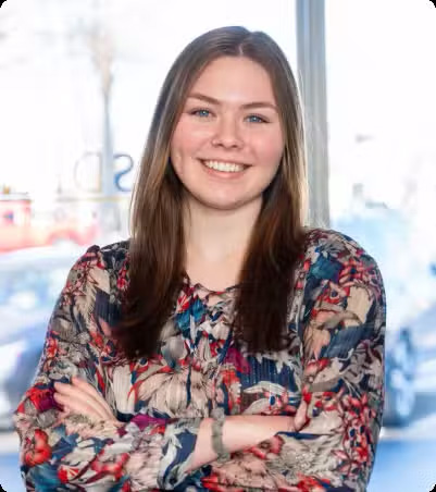 The image shows a smiling woman with short hair, wearing a patterned top, standing against a neutral background, with her arms crossed.