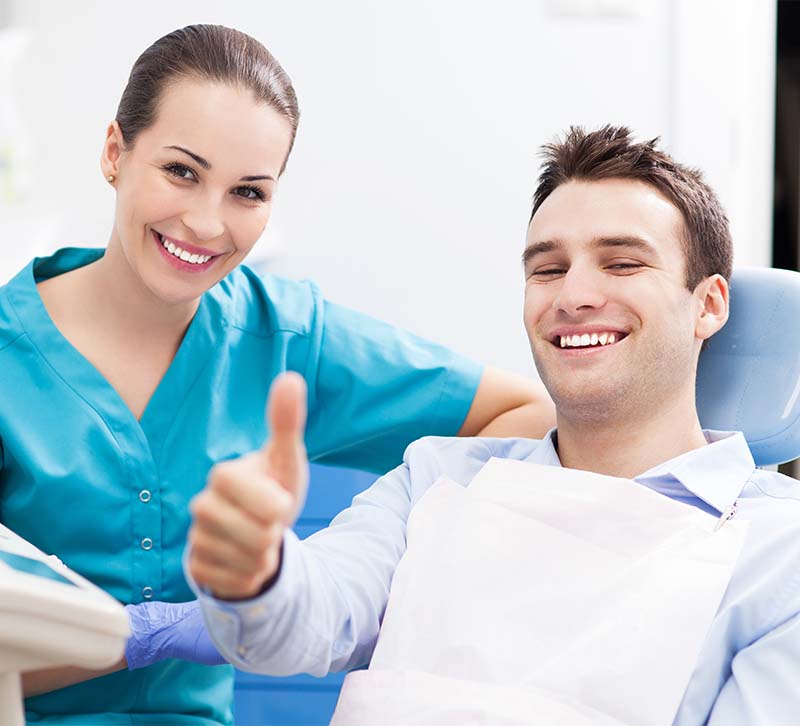 The image shows a dental professional, likely a dentist or dental hygienist, smiling at the camera while seated in front of a patient who is also smiling, both giving thumbs-up signs, suggesting a positive and successful dental visit.