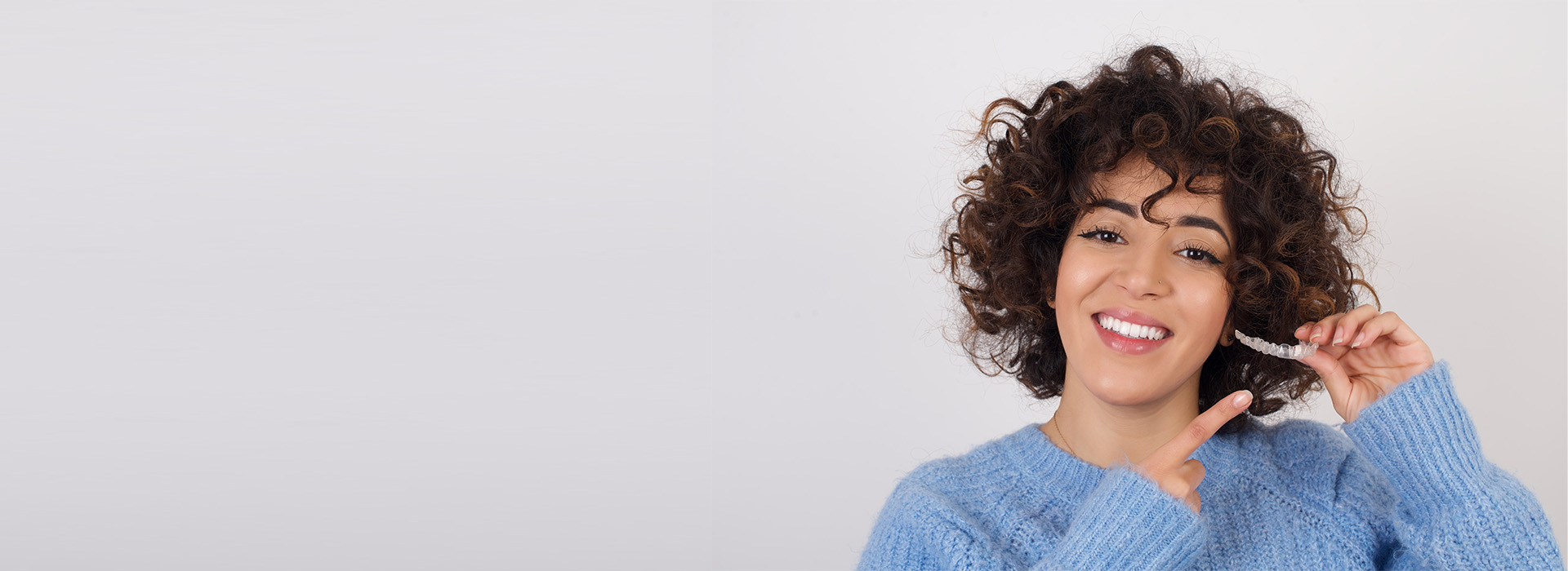 The image shows a person with curly hair smiling at the camera while holding something in their hand. They are wearing a blue sweater and standing against a plain background.