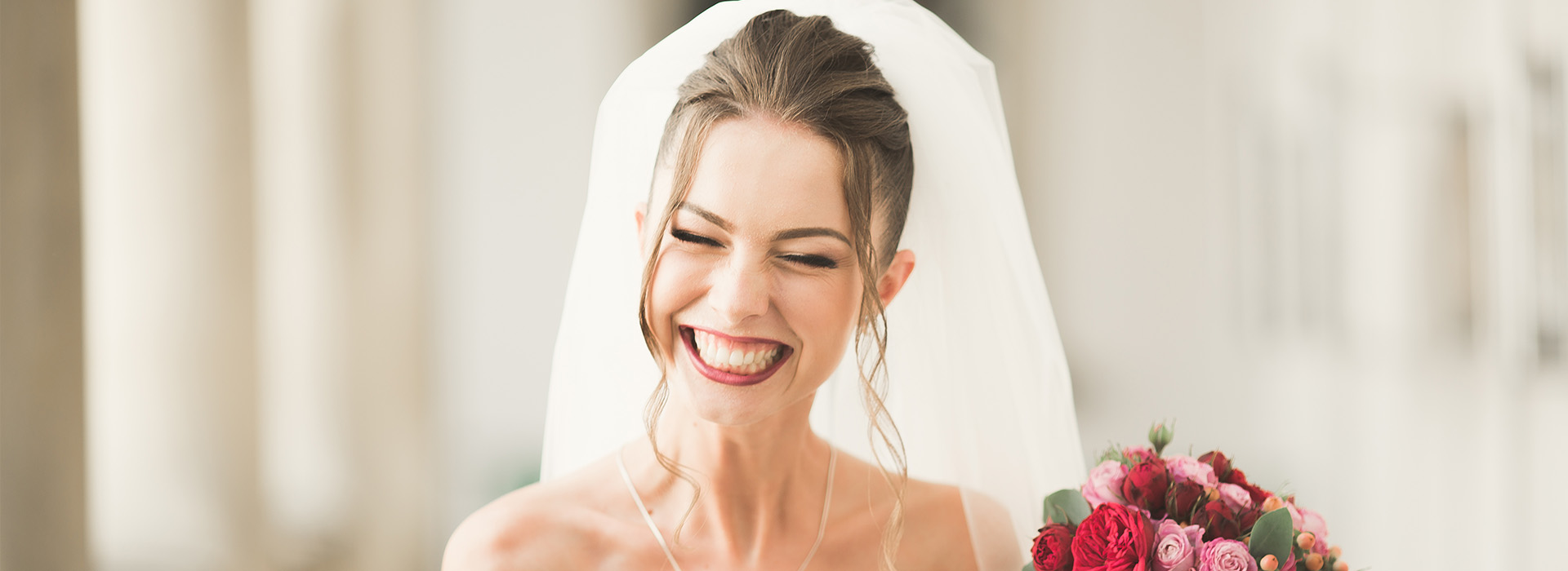 The image shows a woman wearing a white wedding dress with her mouth open in a joyful expression, smiling at the camera, while standing outdoors during the daytime.