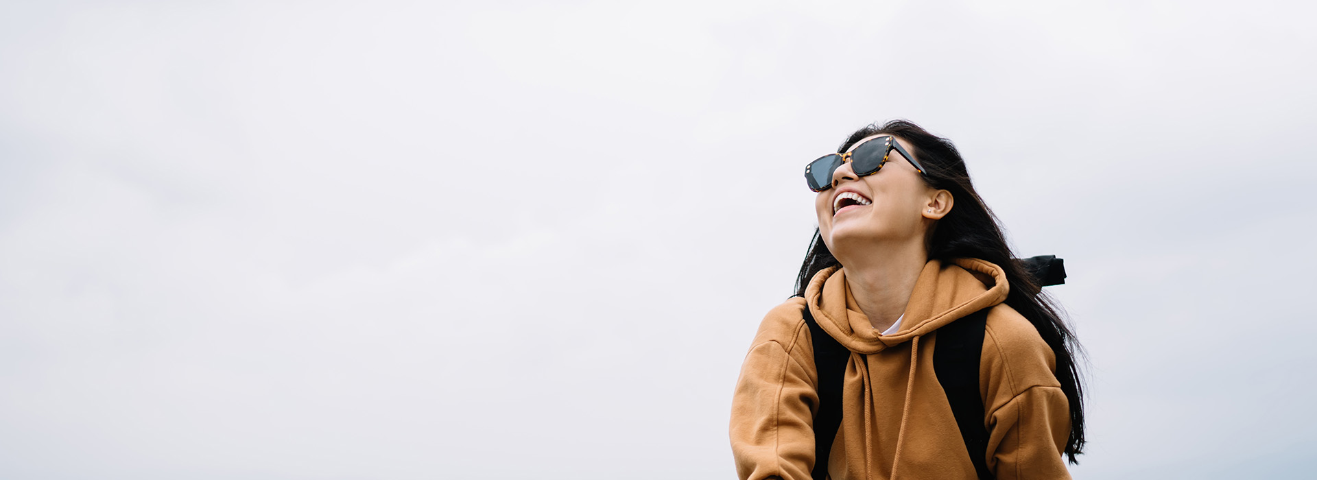 A person stands outdoors with sunglasses, smiling, against a cloudy sky, wearing a backpack and a hooded sweatshirt.