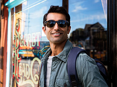 A man wearing sunglasses and a backpack stands outside a shop with a sign visible through the window.