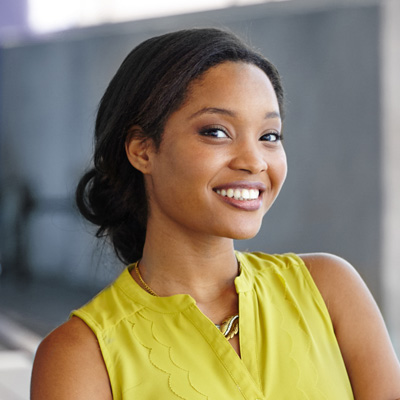 The image shows a smiling woman with dark hair, wearing a yellow top, standing against a backdrop that appears to be an indoor wall.