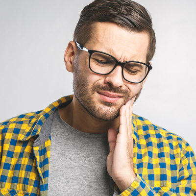 A bearded man with glasses, wearing a blue plaid shirt, stands with his hand on his chin, looking upwards with a concerned expression, possibly in pain.
