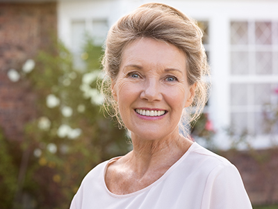 A smiling older woman stands in front of a brick house with white trim, wearing a sleeveless top and her hair styled up.