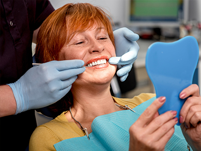 The image depicts a person with red hair sitting in a dental chair, holding a blue dental impression tray on their lap, smiling at the camera while receiving dental care from a professional wearing gloves and a mask.