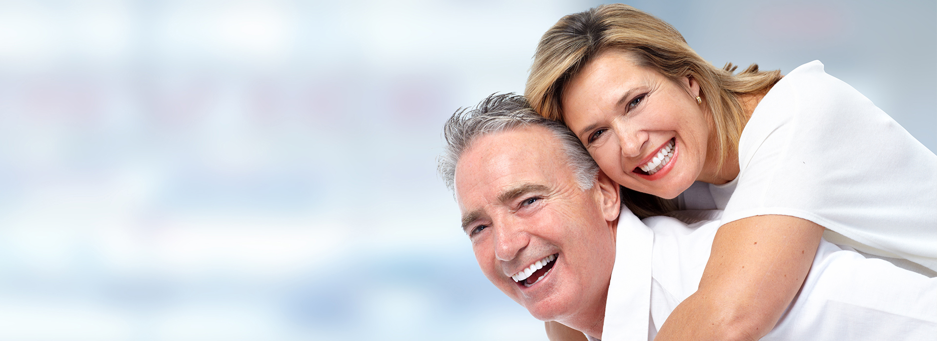 The image depicts two adults, likely a man and a woman, smiling at each other with their arms around one another against a blurred background.