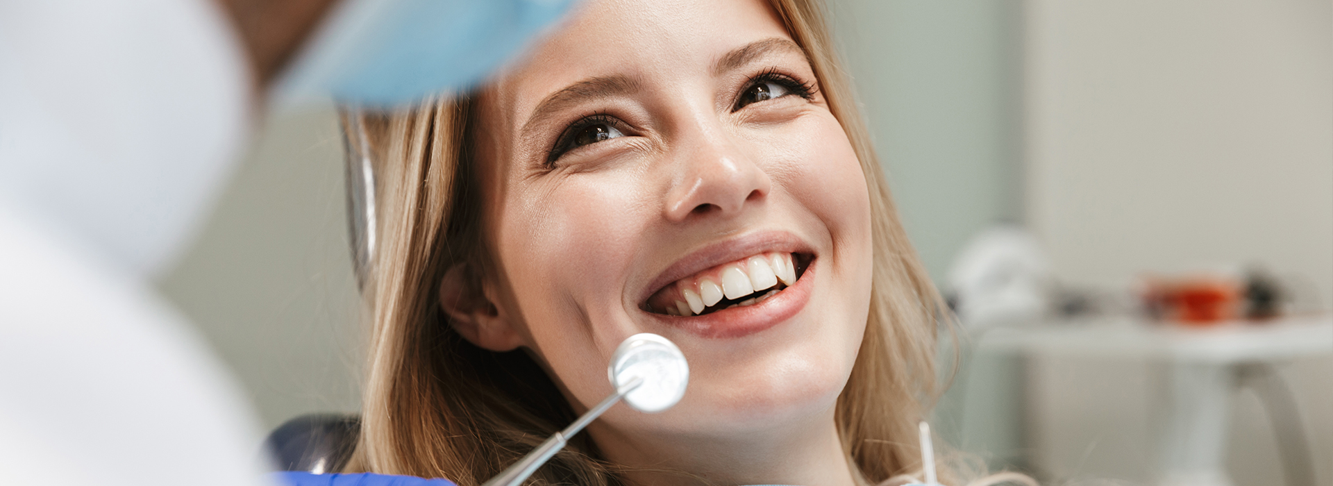 A woman with a bright smile is seated at a dental chair, looking towards the camera, while a dental professional attends to her.