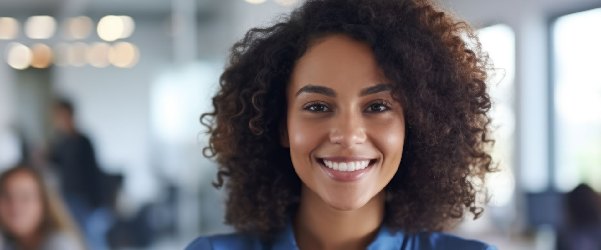 The image shows a woman with short hair smiling at the camera. She appears to be in an office setting, possibly a meeting room, as suggested by the blurred background that includes other people and what looks like a presentation board.