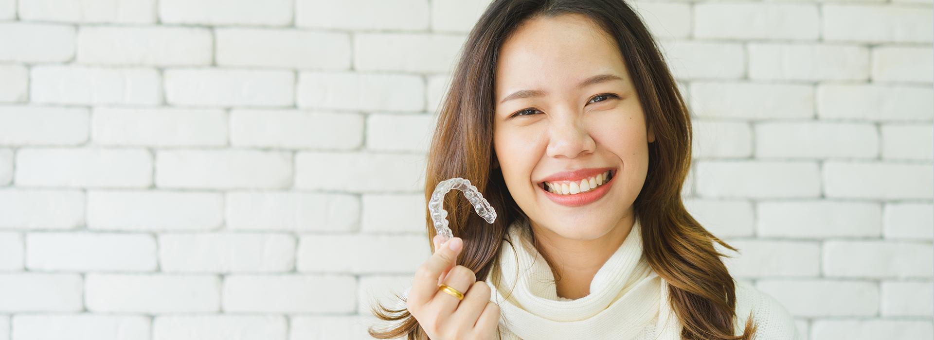 A woman wearing a ring smiles at the camera, set against a brick wall background.