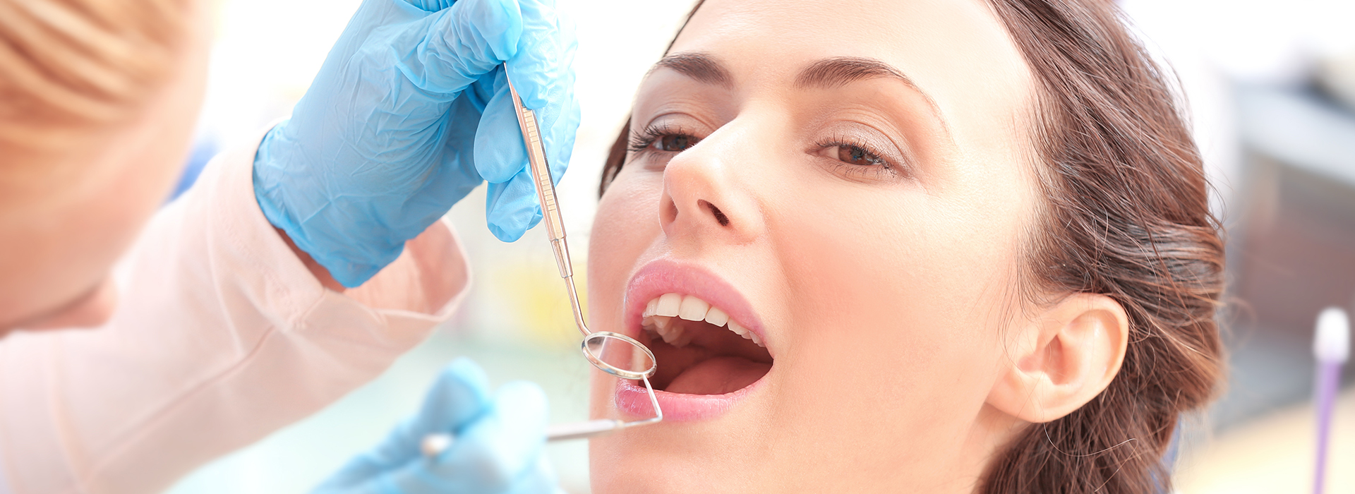 The image shows a woman receiving dental treatment with a dental hygienist performing a cleaning procedure, using dental instruments and wearing protective gloves.