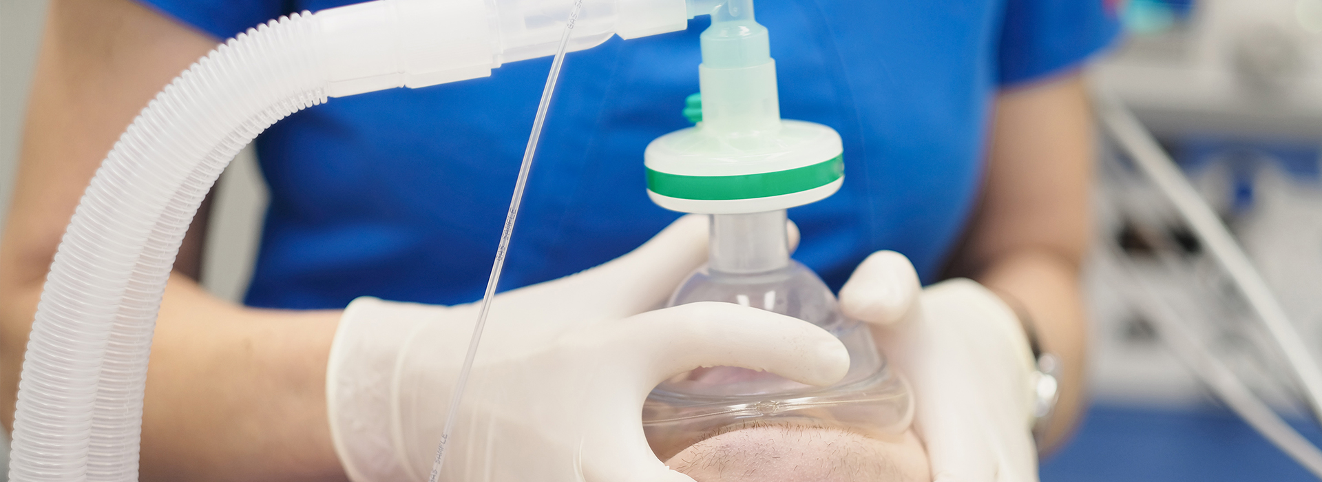 The image shows a person wearing gloves handling an object with a green cap, possibly related to medical equipment or science experiment, set against a blurred background that includes a blue shirt and a section of a white wall.