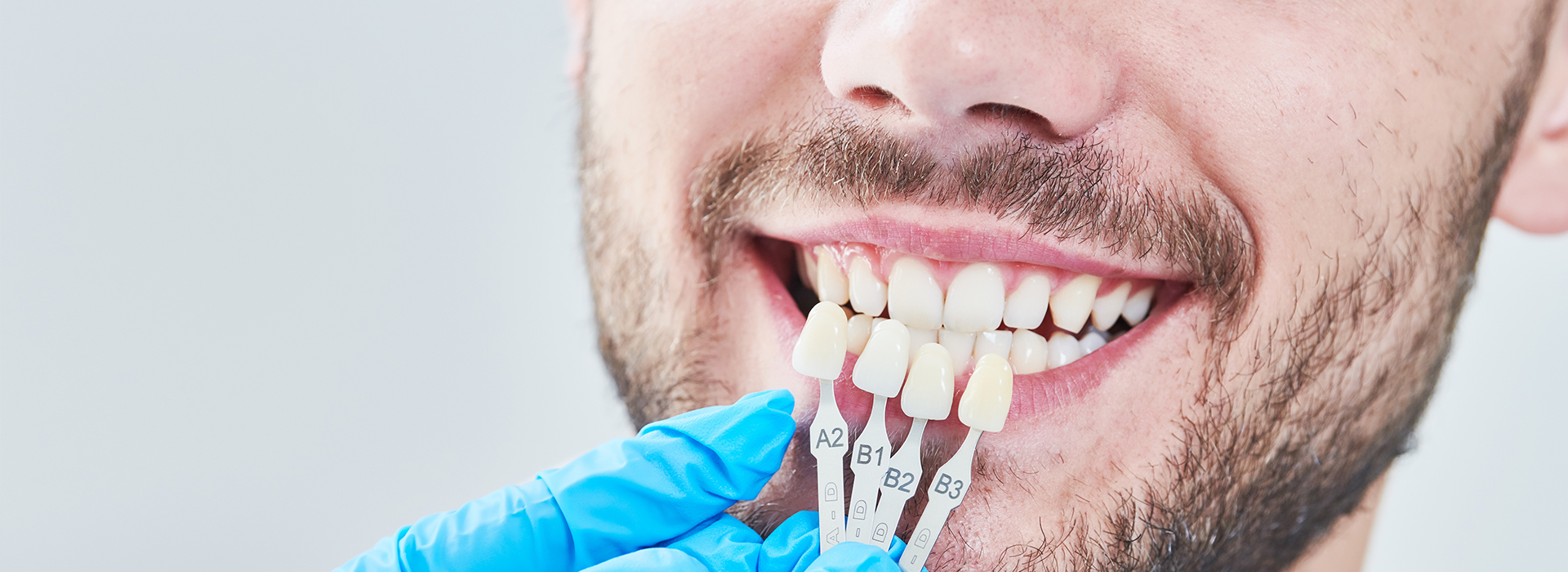 A man with a toothy grin sitting in front of a dental chair, wearing blue gloves and holding a dental instrument, possibly a mirror or probe, near his mouth.