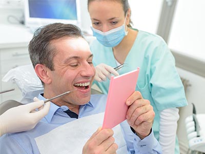 The image shows a man sitting in a dental chair with a smiling expression, looking at a pink card he holds, while being attended to by a female dentist wearing a blue surgical mask and white gloves.