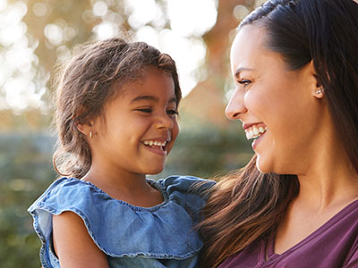 A woman and a young girl are smiling at each other, with the woman holding the child. The setting appears to be an outdoor space, possibly a park, during daylight hours.