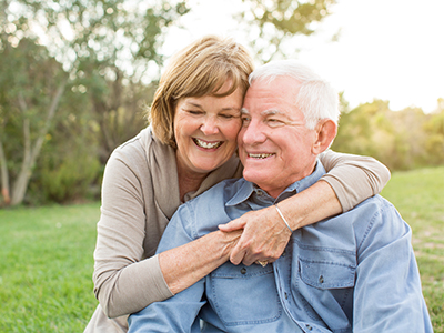 The image shows an elderly couple embracing each other outdoors with a warm smile on their faces, set against a sunset backdrop.