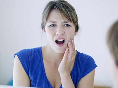 The image shows a woman with her mouth open, expressing surprise or shock, while looking at her reflection in a mirror. She appears to be in an indoor setting with a light blue wall behind her.