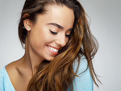 The image shows a smiling woman with long hair, wearing a light blue top, looking towards her left with a slight head tilt.