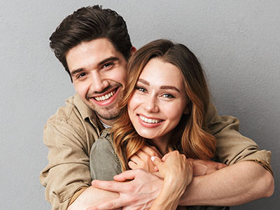 A young couple hugging closely, smiling warmly at each other, with the man wearing a dark shirt and the woman in a light-colored top.