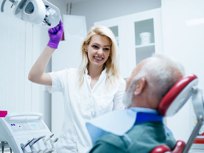 A dental hygienist in a white coat attends to an elderly man with white hair seated in a dentist s chair, holding a mirror while smiling.