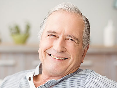 The image shows a smiling older man with gray hair, wearing glasses, sitting comfortably indoors with his eyes closed and hands resting on his chest.