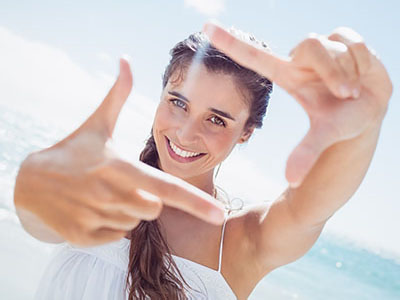 A woman with long hair is taking a selfie at the beach, smiling and holding up her hand with fingers extended towards the camera.